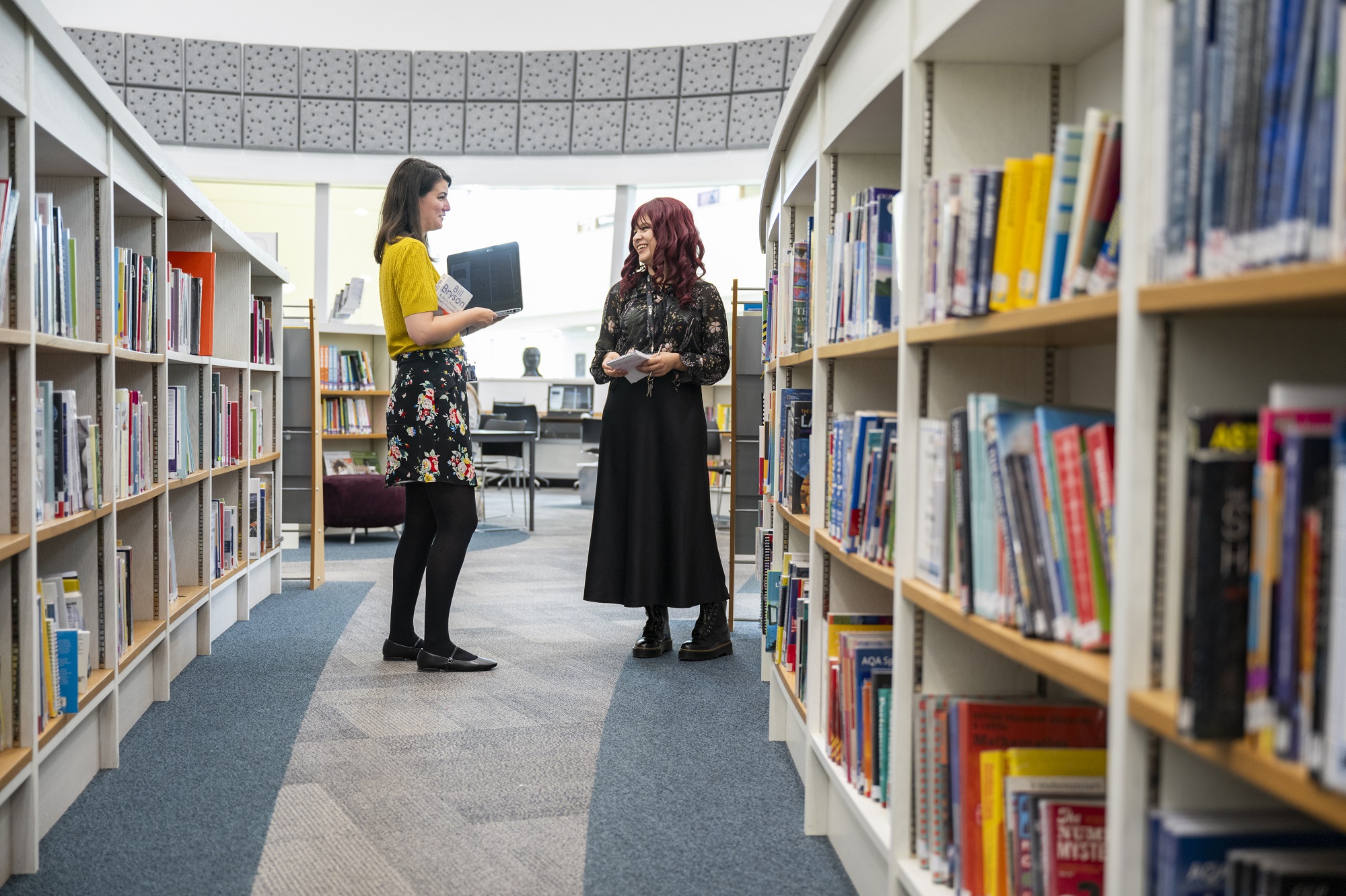 Two ladies chatting in a library - demonstrating a engaging and supportive environment for those who choose to work for us.